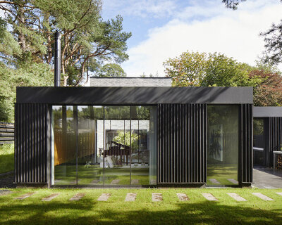 a black garden pavilion wraps around a two-story brick house in a leafy scottish suburb