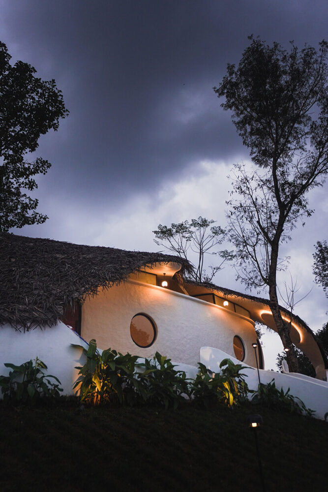 the retreat glows softly against the Wayanad sky, highlighting its curving shell walls and thatched roof