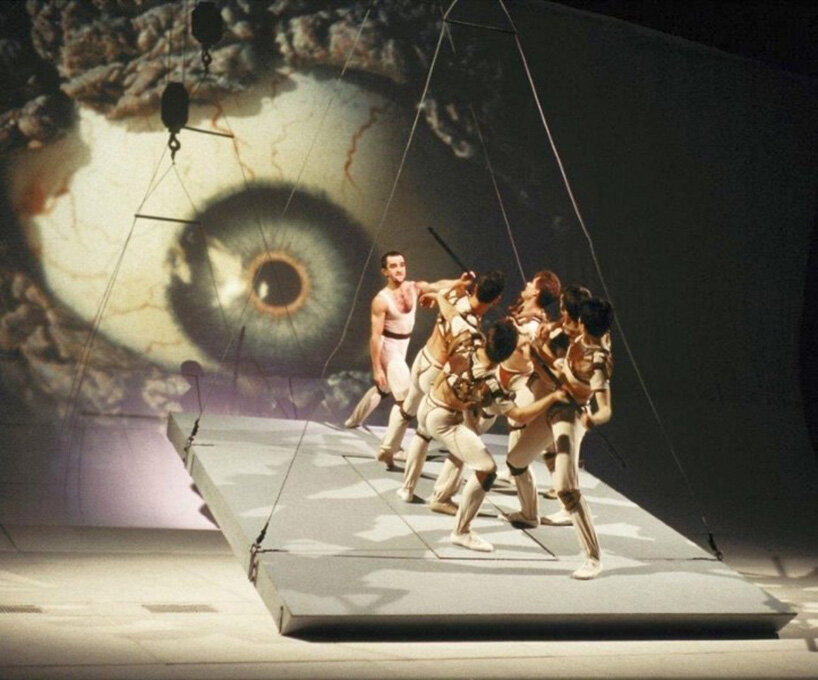 dancers perform on a raised platform beneath a large projected eye